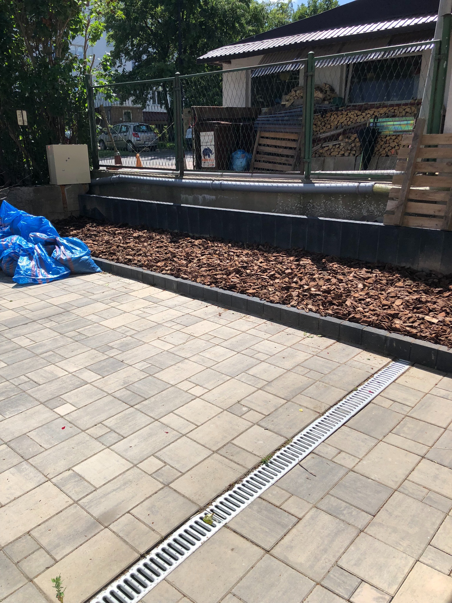 Wykończony fragment ogrodu z brukowanym podjazdem, obrzeżem z kory i drenażem liniowym. W tle widoczna siatka ogrodzeniowa i składowane drewno.