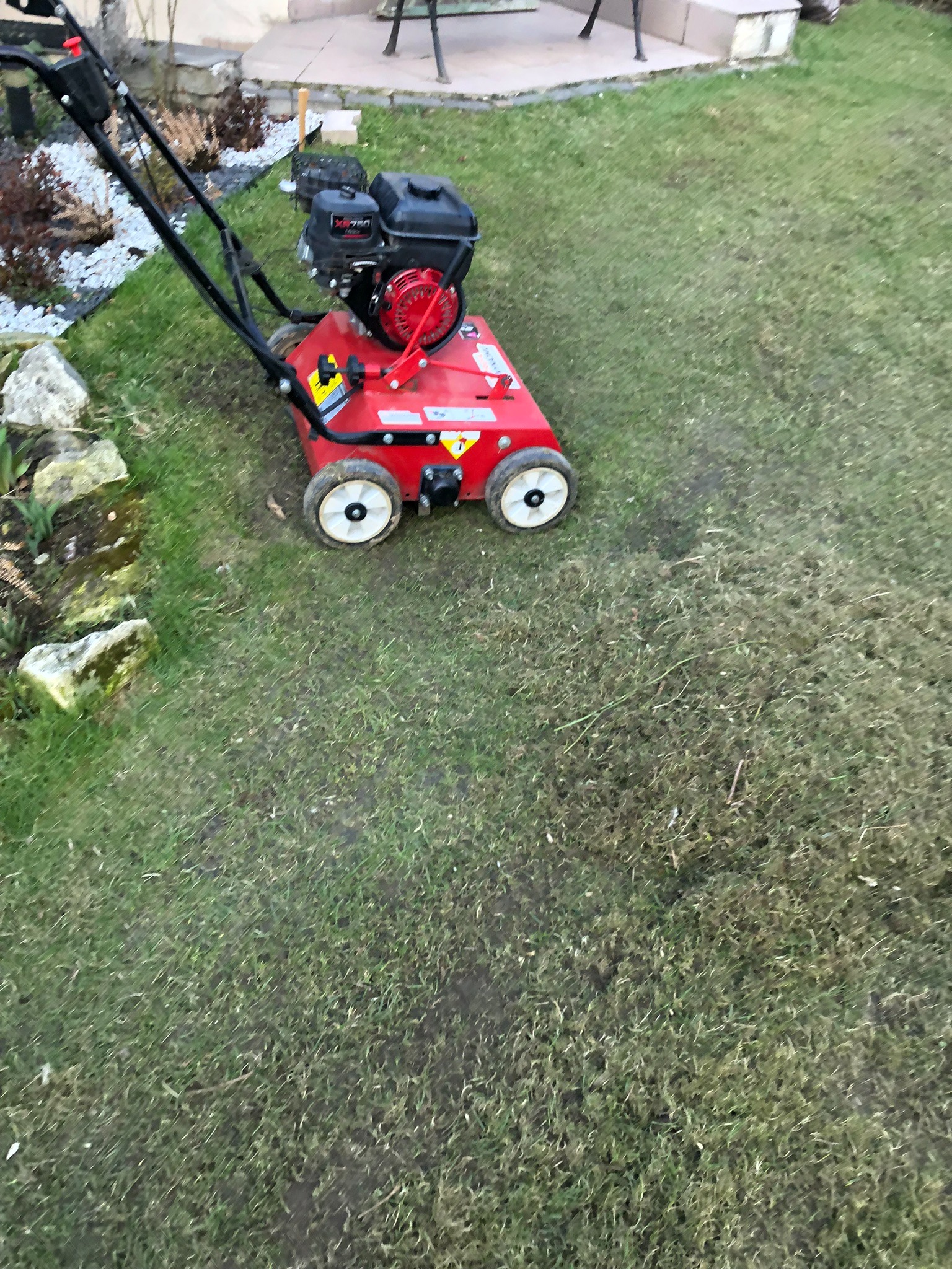 Aerator spalinowy na trawniku. Widoczne białe koła i czerwona obudowa urządzenia. Trawa miejscami przerzedzona, w tle elementy ogrodu i siedzisko.