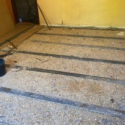 FS Tech - Montaż, remonty, elektryka - Przygotowanie podłoża pod podłogę: legary z przestrzenią wypełnioną wiórami. Widoczny fragment ściany i narzędzia. Etap prac remontowych w pomieszczeniu o żółtych ścianach.