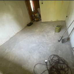 FS Tech - Montaż, remonty, elektryka - Wnętrze pokoju w trakcie remontu. Świeżo wylana wylewka podłogowa, widoczne narzędzia: szpachla, łopata, przewód zasilający. Tło stanowią ściany w stanie surowym.