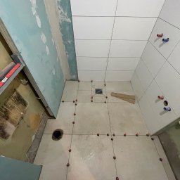 FS Tech - Montaż, remonty, elektryka - Białe płytki ułożone na podłodze i ścianie łazienki w trakcie remontu. Widoczne krzyżyki dystansowe i fragmenty ścian z płytami kartonowo-gipsowymi.