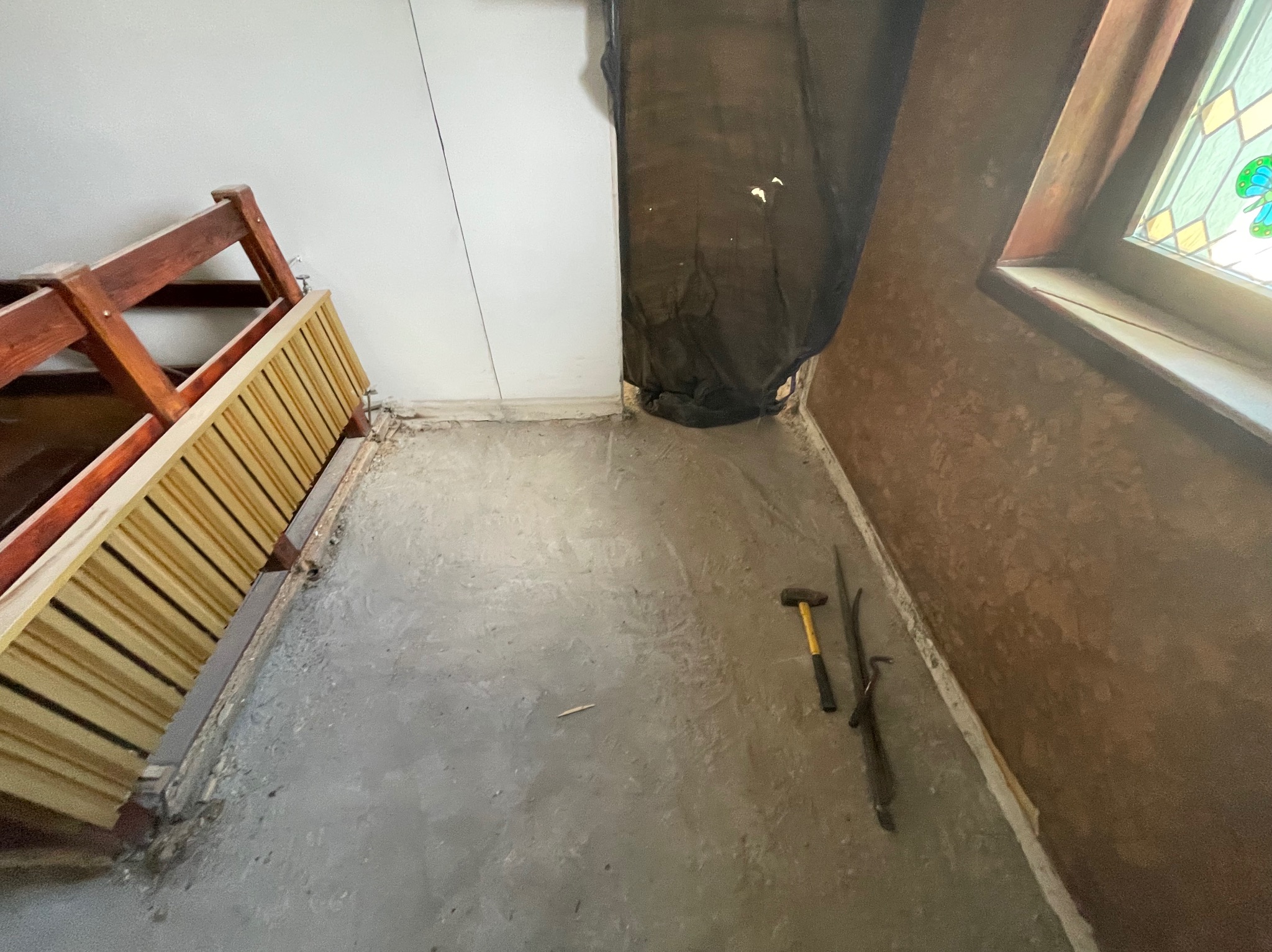 Przygotowanie do remontu: fragment pokoju z odsłoniętą podłogą, narzędzia (młotek) leżące obok ściany. Widoczna część drewnianej konstrukcji i okno ze szkłem witrażowym.