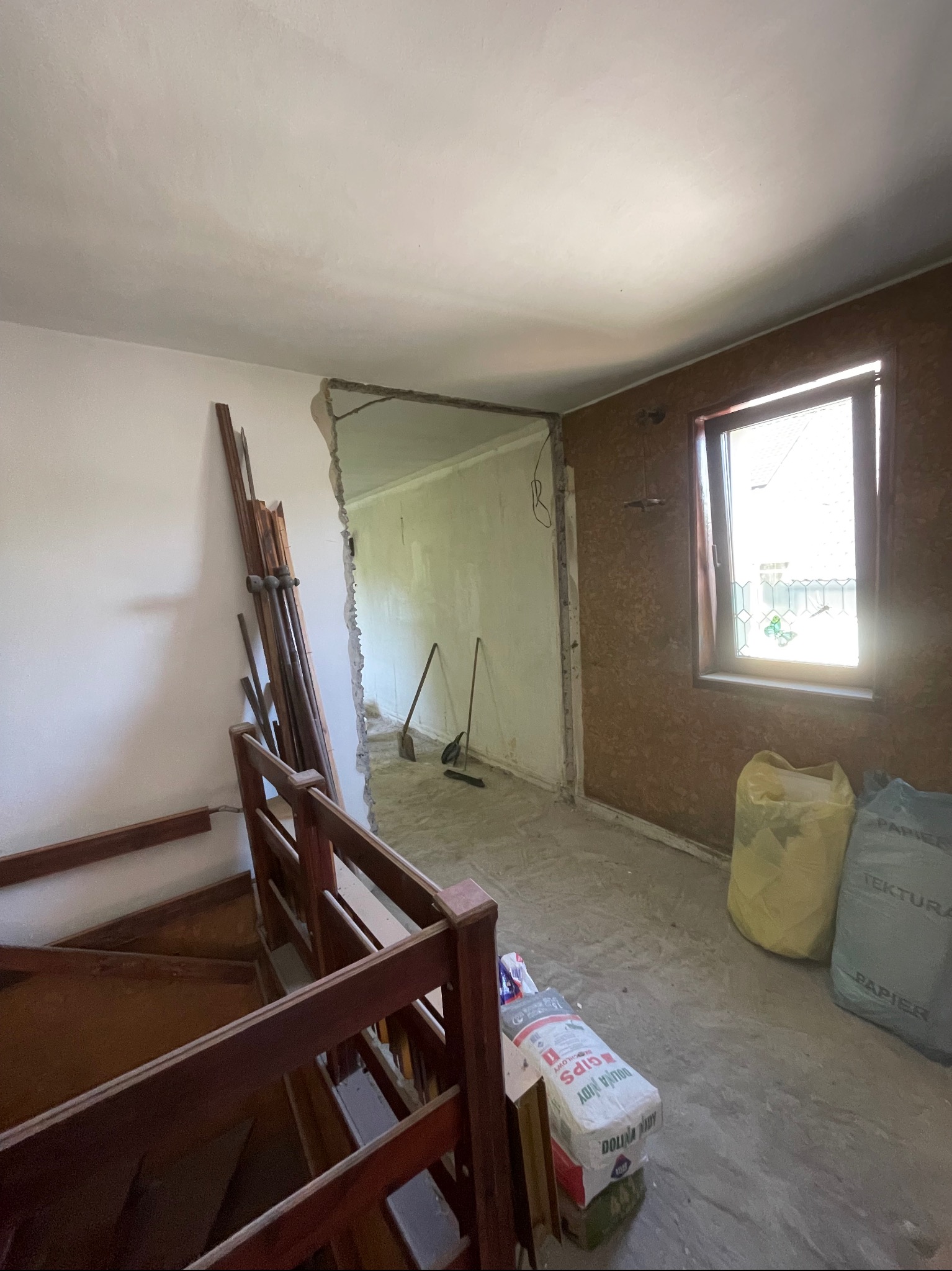 Wnętrze w trakcie remontu: widoczne worki z gipsem, usunięta część ściany, drewniane schody i okno z widokiem na zewnątrz. Etap przygotowania do dalszych prac wykończeniowych.