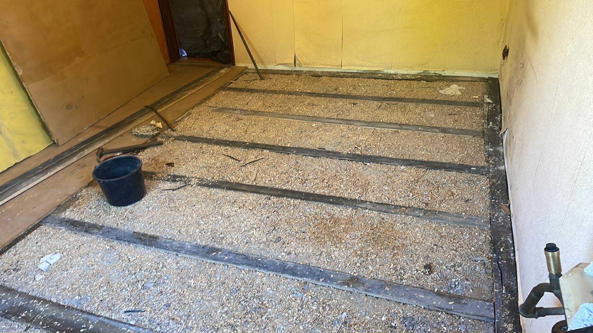 Przygotowanie podłoża pod podłogę: legary z przestrzenią wypełnioną wiórami. Widoczny fragment ściany i narzędzia. Etap prac remontowych w pomieszczeniu o żółtych ścianach.