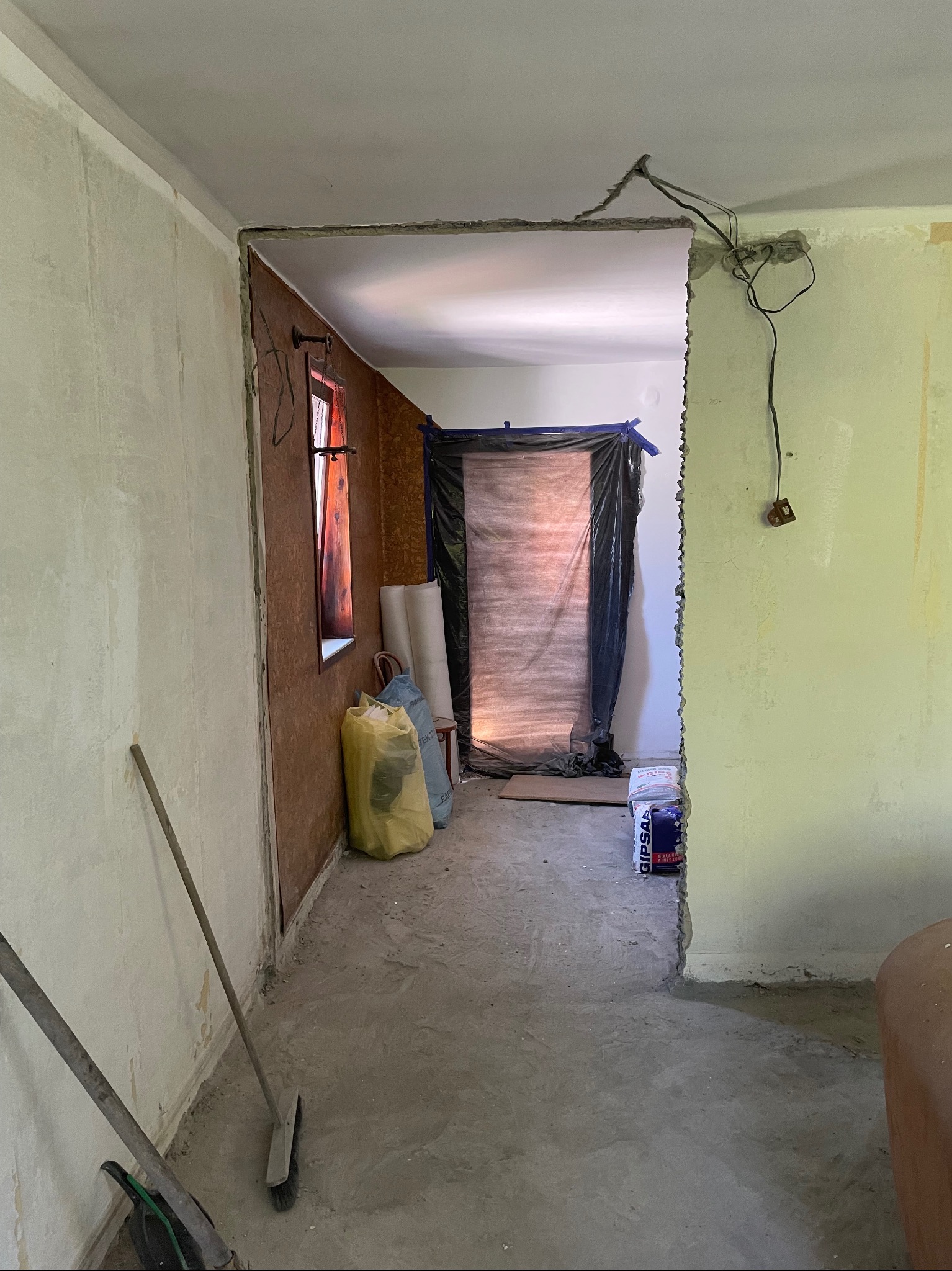 Wnętrze w trakcie remontu: odsłonięte przewody elektryczne, worki z materiałami budowlanymi, ściany przygotowane do gładzi, widoczne narzędzia i zabezpieczone folią przejście.