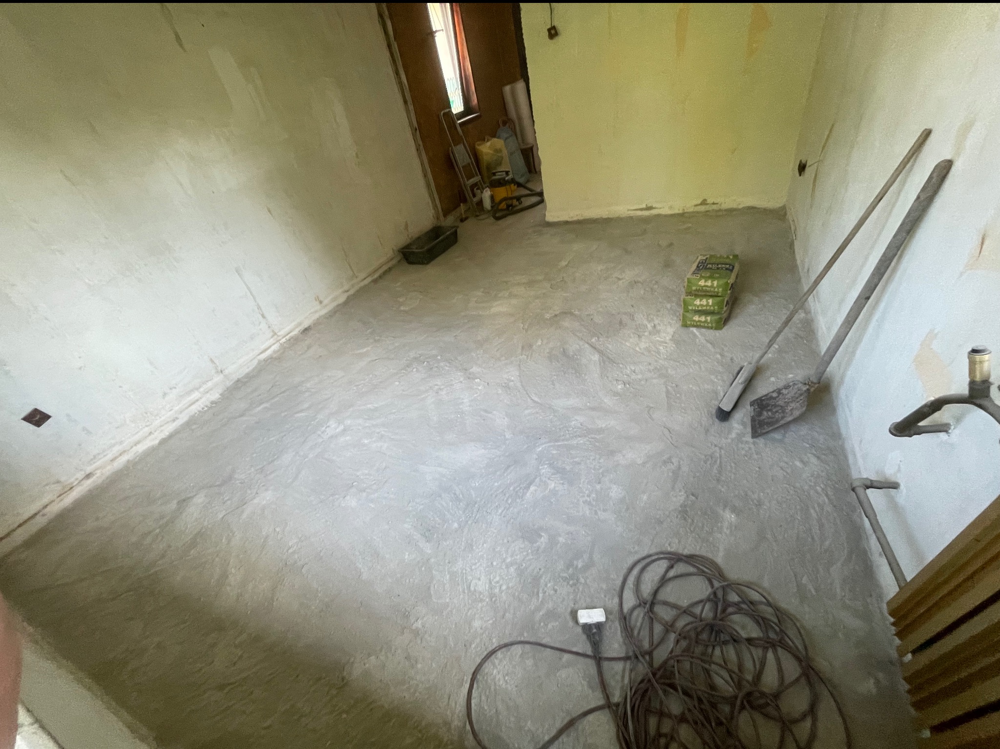 Wnętrze pokoju w trakcie remontu. Świeżo wylana wylewka podłogowa, widoczne narzędzia: szpachla, łopata, przewód zasilający. Tło stanowią ściany w stanie surowym.