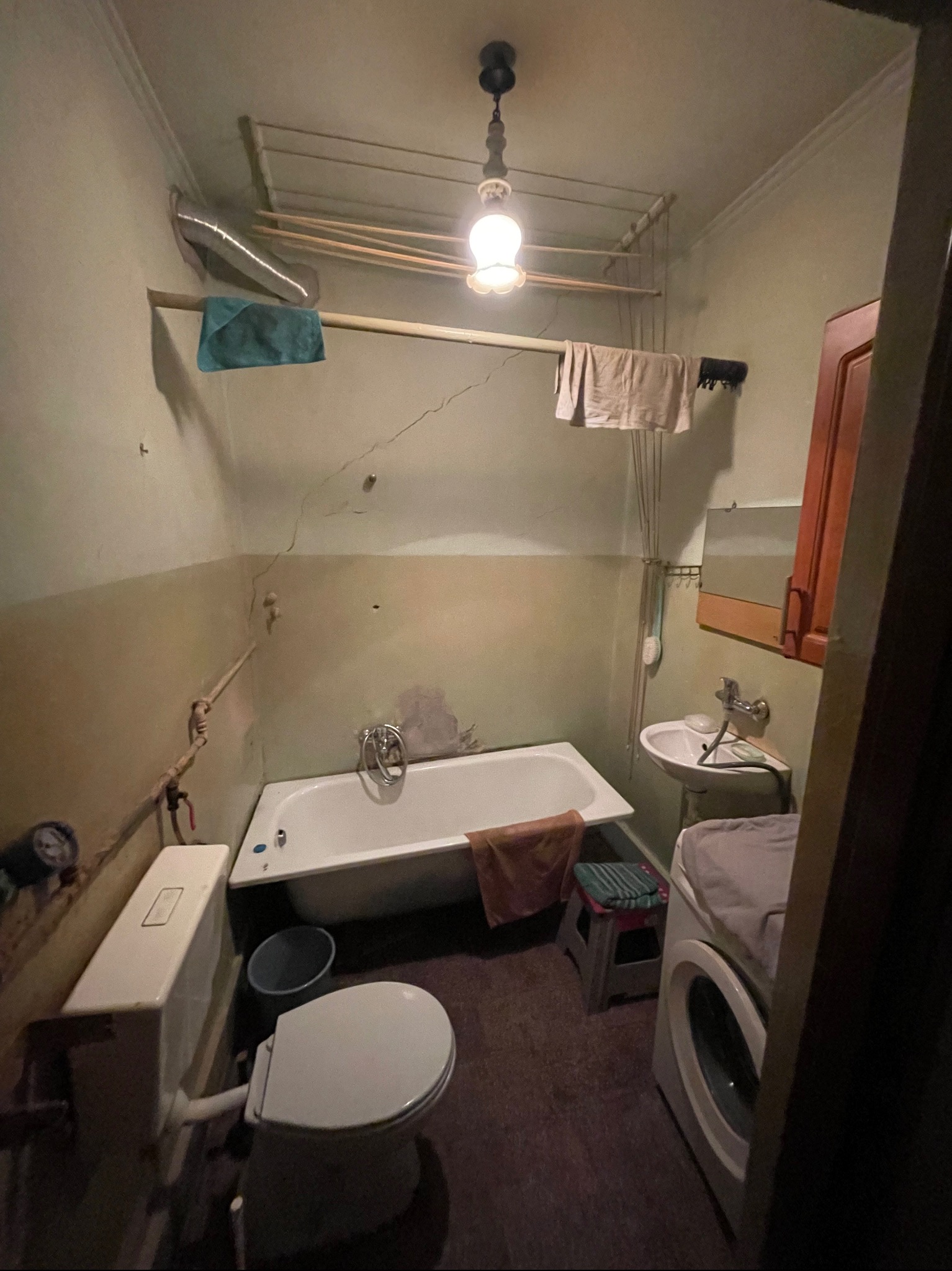 Zdjęcie wnętrza łazienki z wanną, umywalką, toaletą i pralką. Widoczne pęknięcia na ścianach, suszarka pod sufitem i wiszące ręczniki. Oświetlenie centralne.