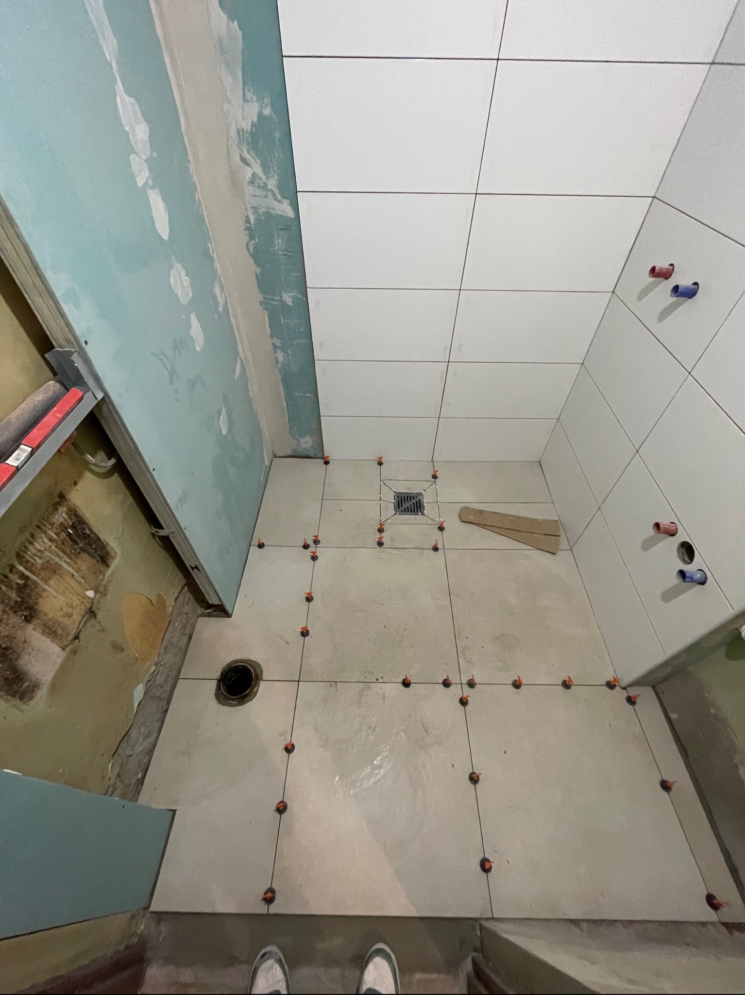 Białe płytki ułożone na podłodze i ścianie łazienki w trakcie remontu. Widoczne krzyżyki dystansowe i fragmenty ścian z płytami kartonowo-gipsowymi.