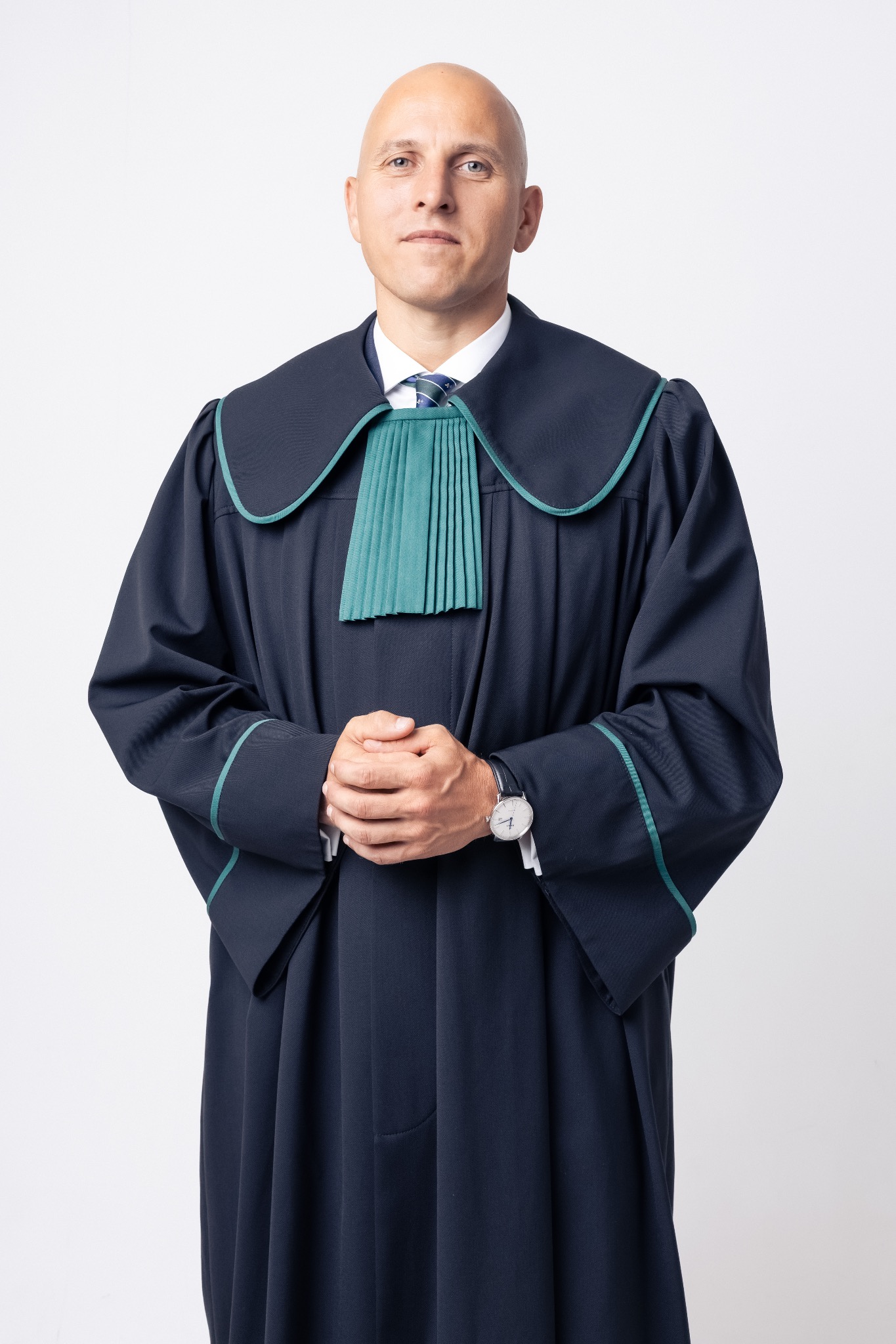 Portret mężczyzny w todze adwokackiej na jasnym tle, z rękami złożonymi przed sobą i widocznym zegarkiem na nadgarstku. Ubiór galowy z zielonymi akcentami.