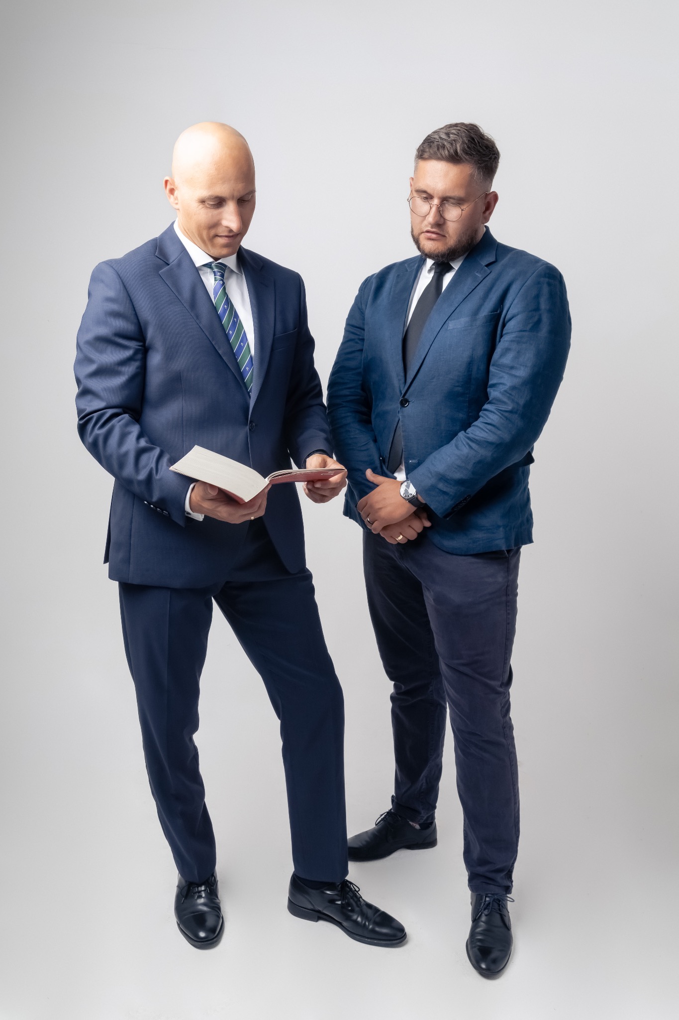 Dwóch prawników w garniturach analizuje dokumenty na neutralnym tle, jeden z nich w okularach, obaj w pozycji stojącej, z widocznymi butami i częścią nóg.