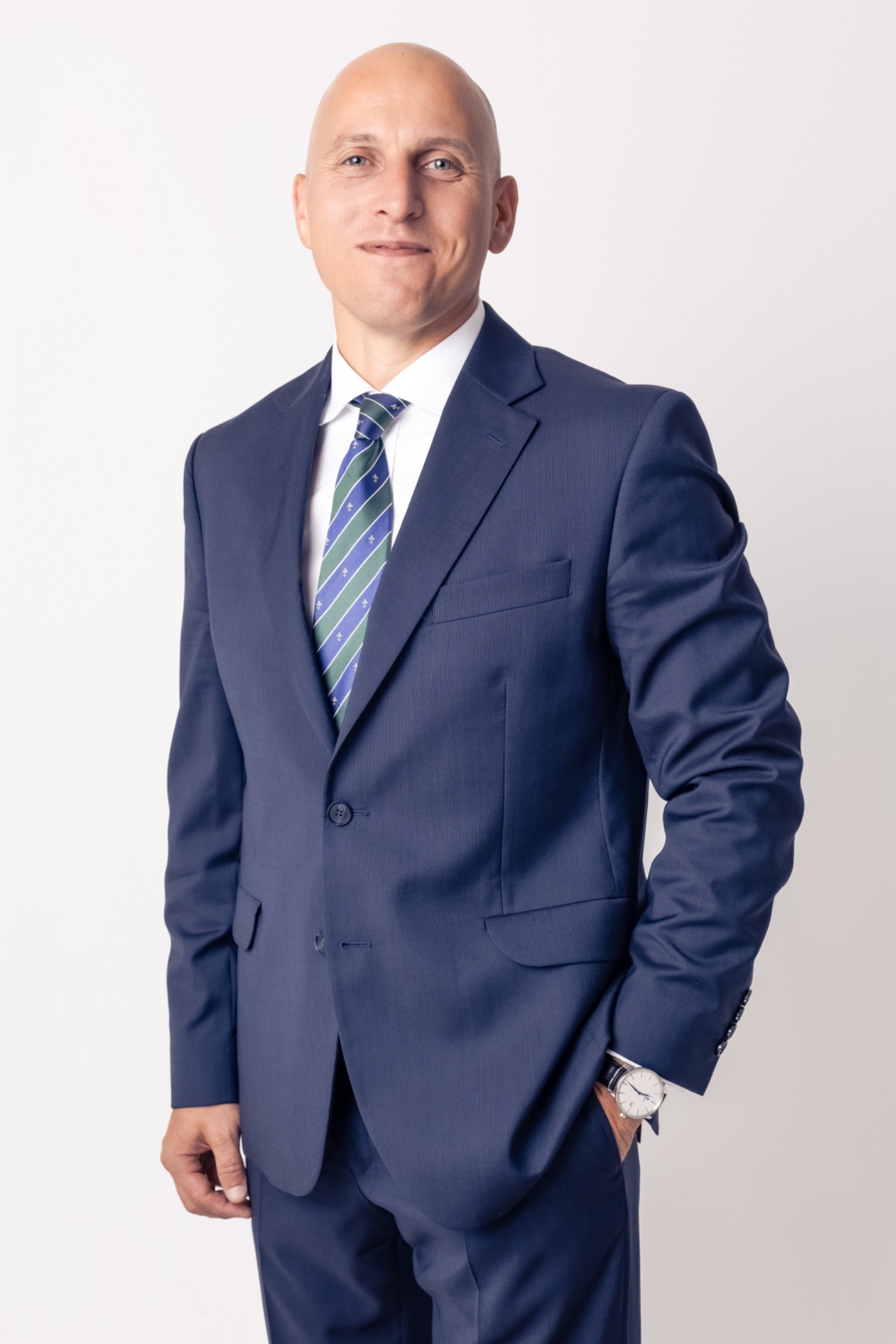 Portret mężczyzny w granatowym garniturze, białej koszuli i krawacie w niebiesko-zielone pasy, z zegarkiem na ręku, pozującego na jasnym tle.