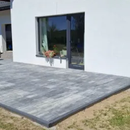 Szara kostka brukowa ułożona na tarasie przylegającym do białego budynku z oknem, na którym stoją kwiaty. Z boku zaparkowany srebrny samochód.