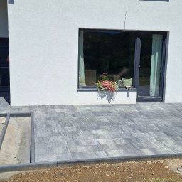 Bager Kop - Ułożona szara kostka brukowa przed nowoczesnym domem z białymi ścianami. Widoczny fragment podjazdu z szarego żwiru i skrzynka z owocami.