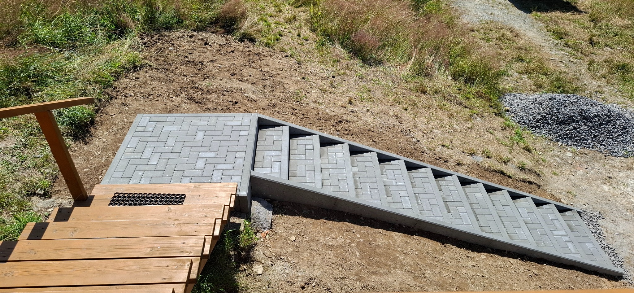 Schody z kostki brukowej prowadzące z drewnianego tarasu na zbocze, z widocznym stosem żwiru i wykończeniem terenu w tle. Jasne, naturalne oświetlenie.