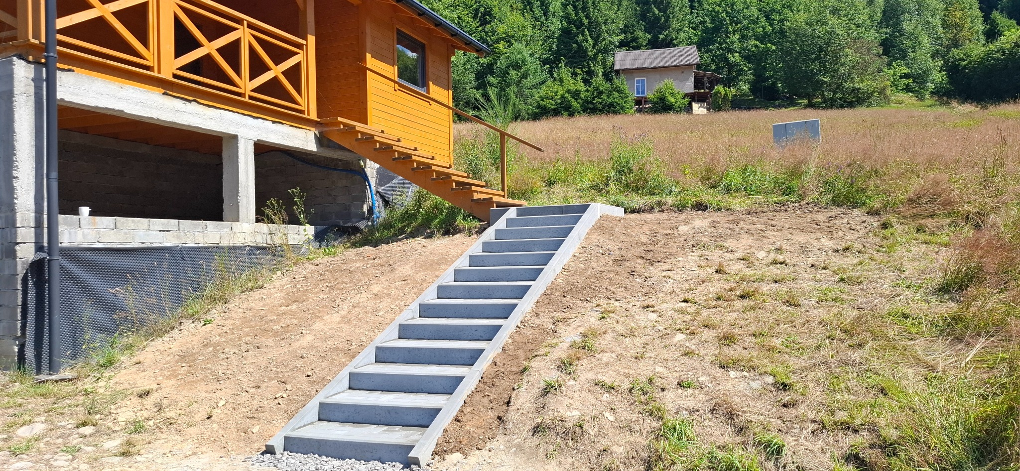 Betonowe schody prowadzą od domu na wzgórzu, z widocznym drewnianym domkiem w tle, otoczone zielenią i ziemią.