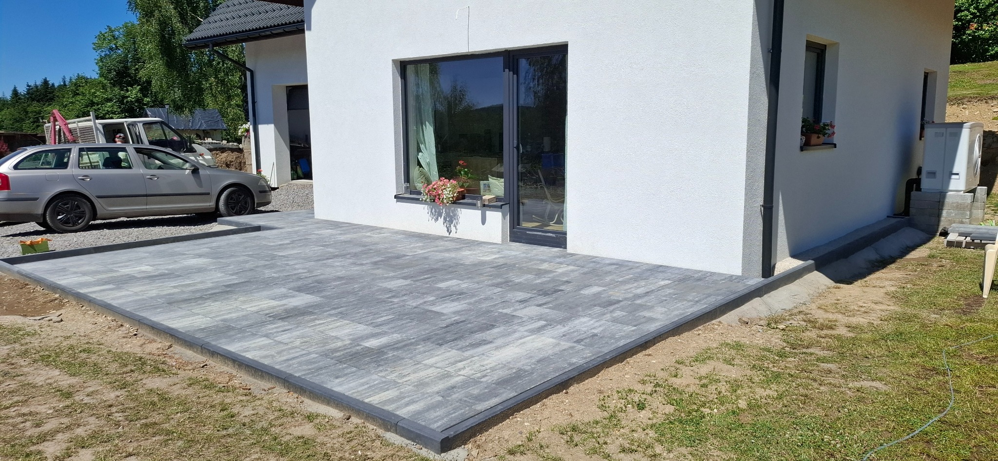 Szara kostka brukowa ułożona na tarasie przylegającym do białego budynku z oknem, na którym stoją kwiaty. Z boku zaparkowany srebrny samochód.