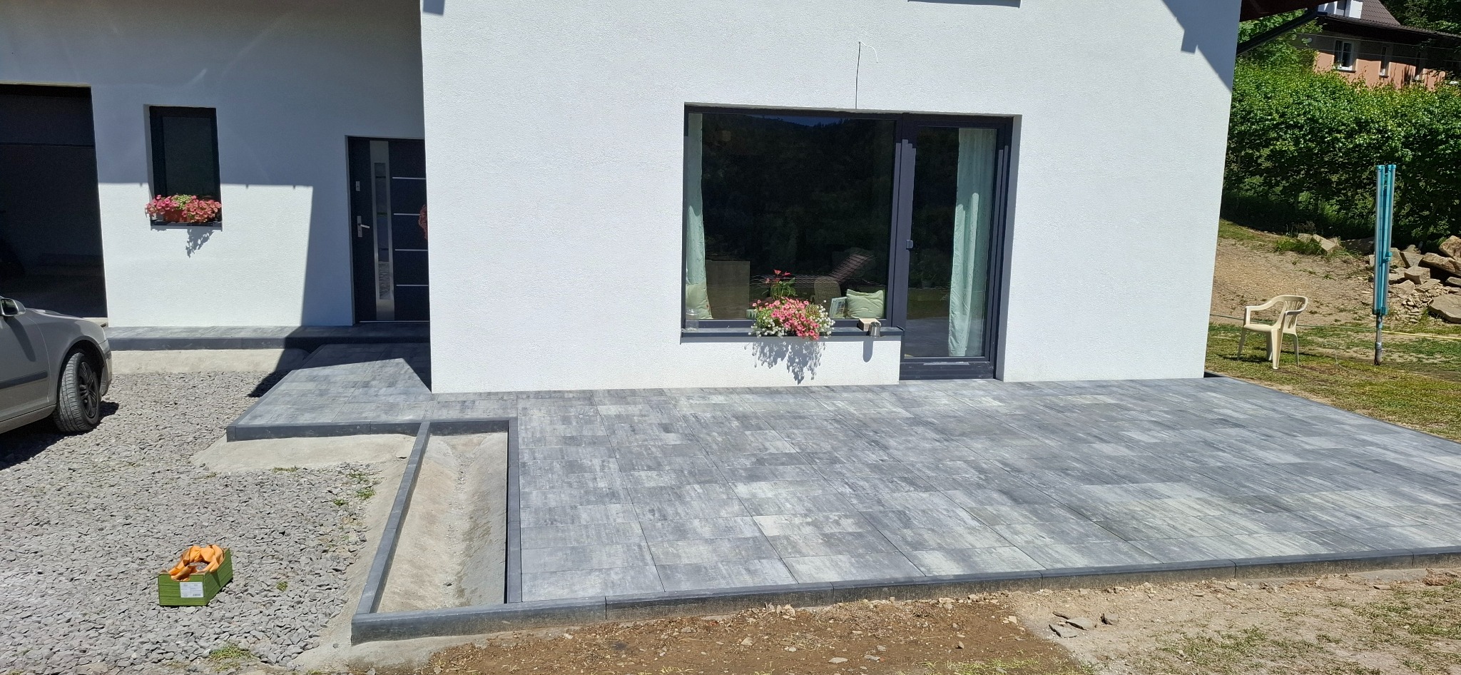 Ułożona szara kostka brukowa przed nowoczesnym domem z białymi ścianami. Widoczny fragment podjazdu z szarego żwiru i skrzynka z owocami.