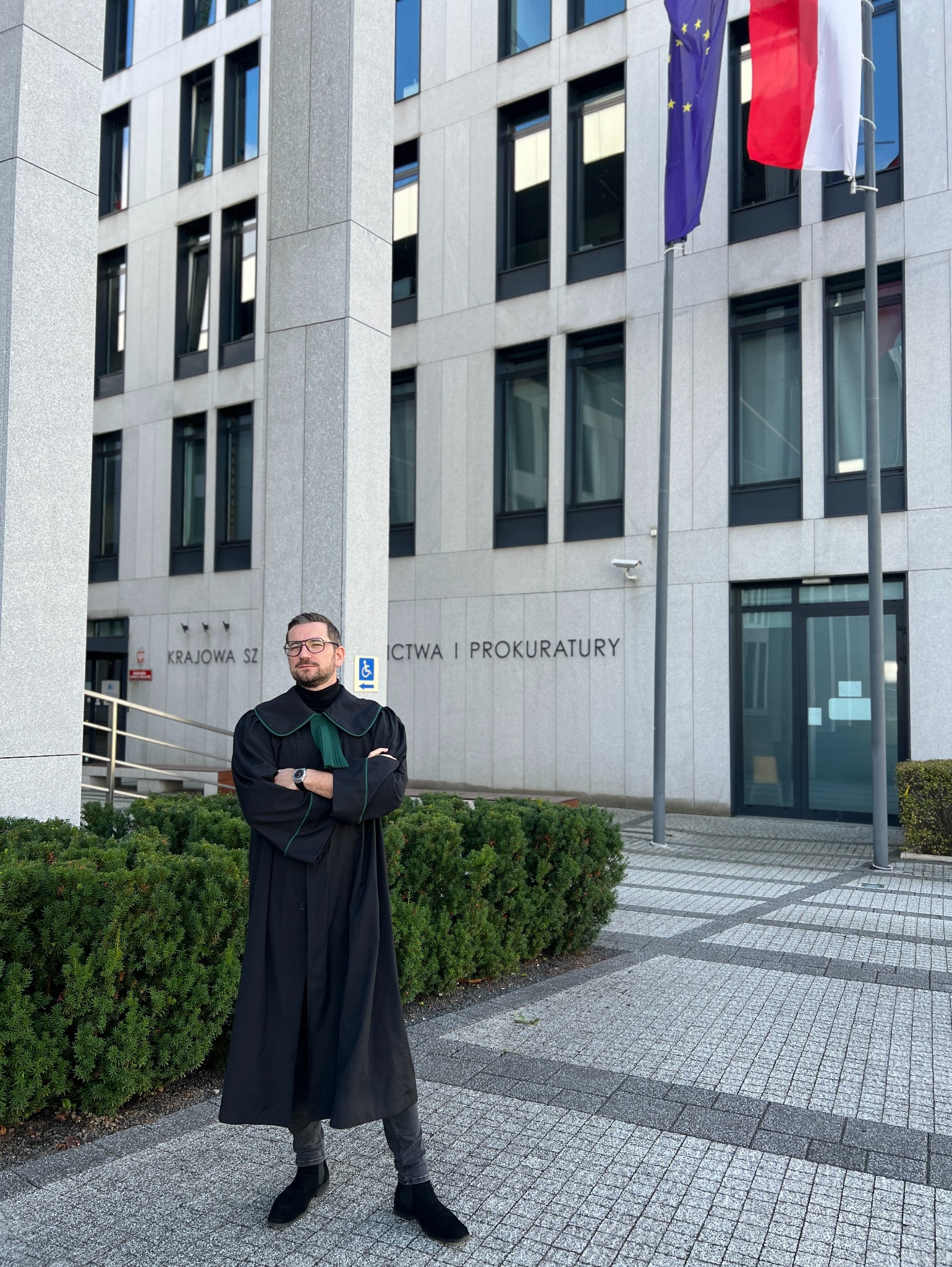 Adwokat w todze przed budynkiem Krajowej Szkoły Sądownictwa i Prokuratury. Widoczne flagi Polski i UE. Osoba z założonymi rękami, profesjonalny wygląd.
