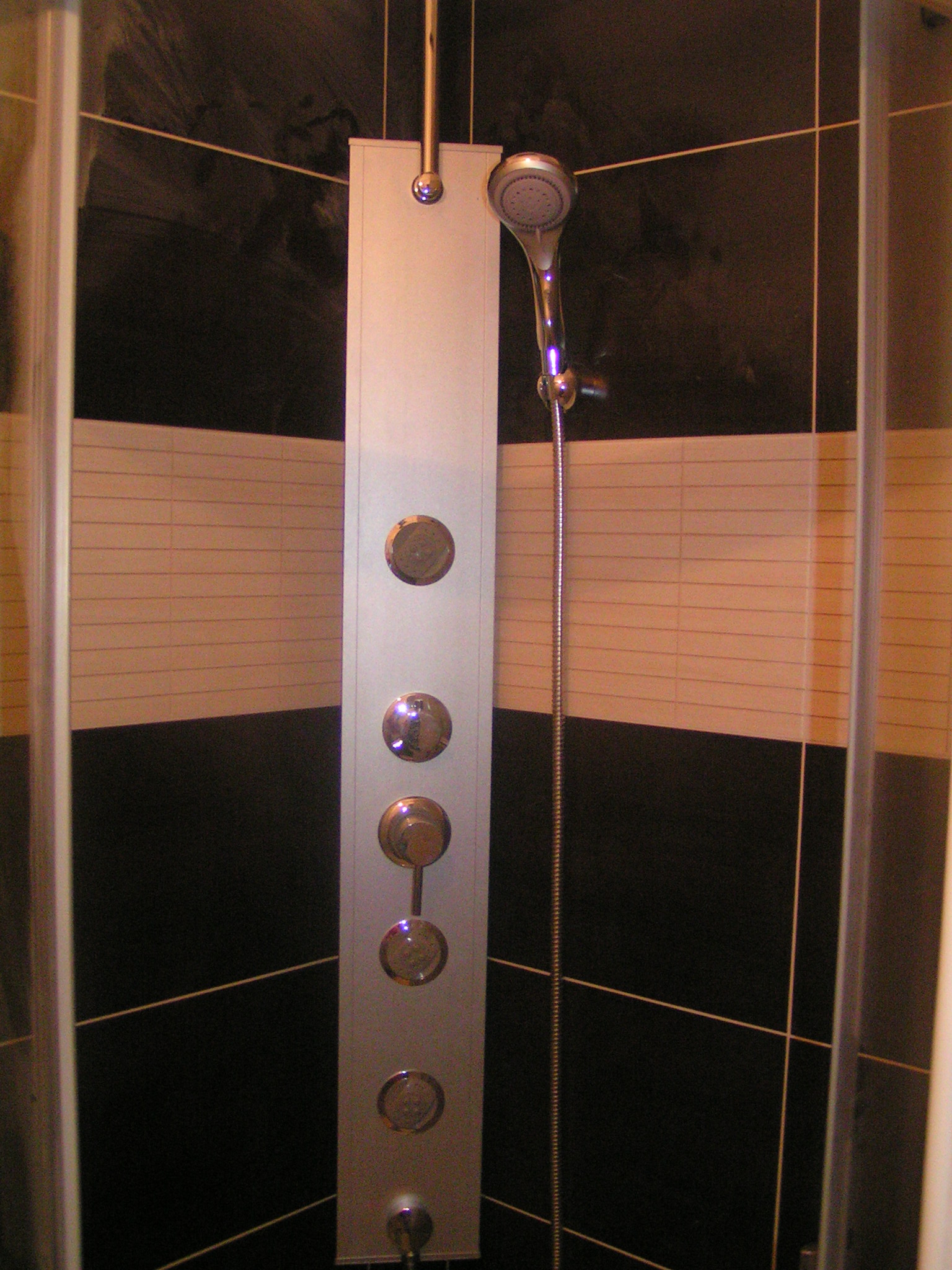 Panel prysznicowy z dyszami i słuchawką, zamontowany na ścianie z czarno-białych płytek w kabinie prysznicowej.