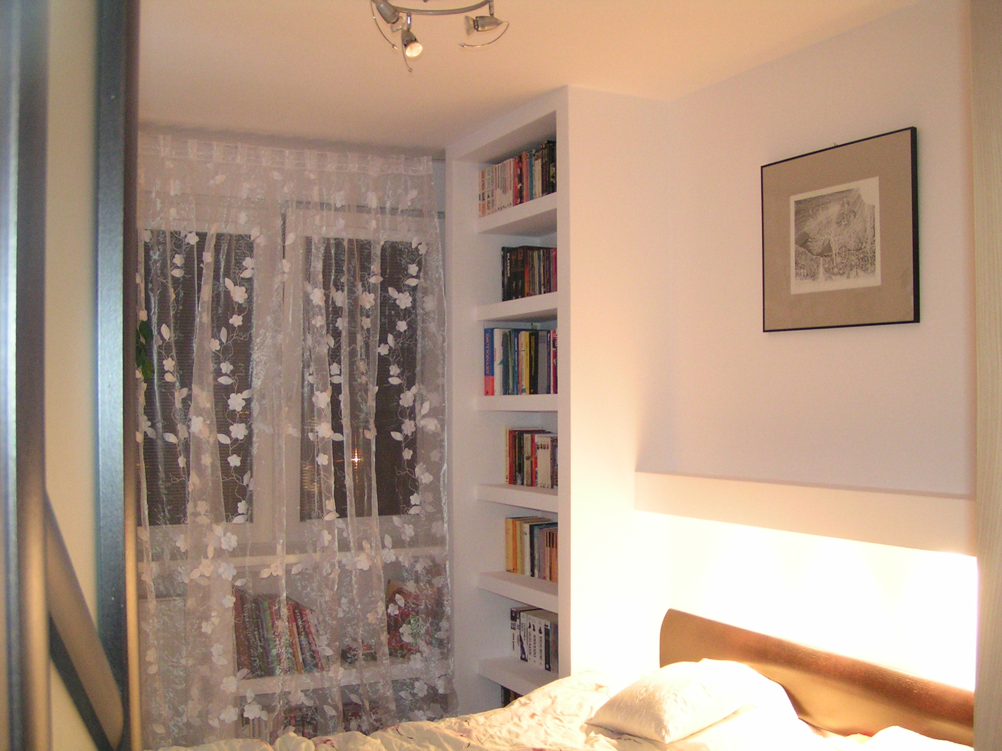 Jasne wnętrze sypialni z wbudowanym regałem na książki, firankami w kwiaty i obrazem w ramie nad wezgłowiem łóżka.