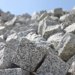 Stos szarej kostki granitowej w surowym stanie pod błękitnym niebem. Widoczna faktura kamienia i nieregularne kształty kostek. Ujęcie z dołu, podkreślające ilość materiału.