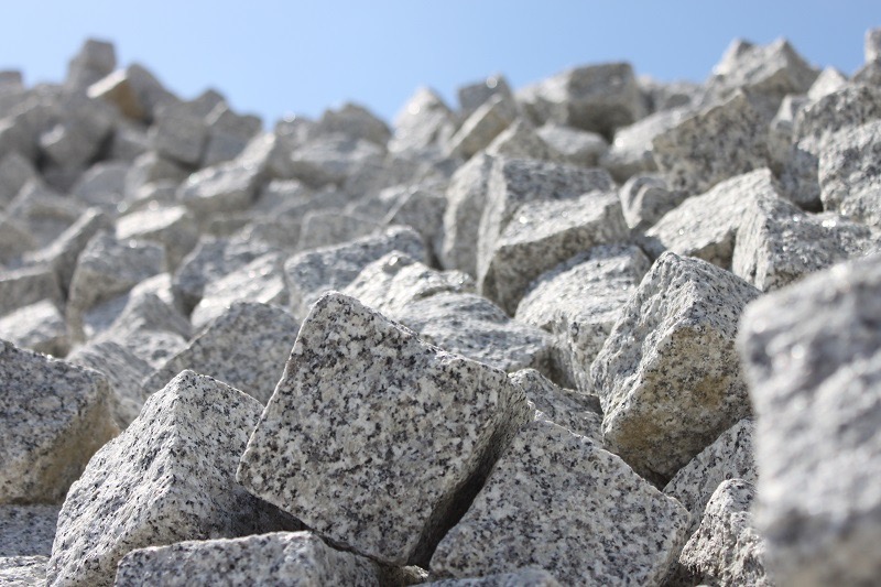 Stos szarej kostki granitowej w surowym stanie pod błękitnym niebem. Widoczna faktura kamienia i nieregularne kształty kostek. Ujęcie z dołu, podkreślające ilość materiału.