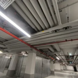 Instalacja rur pod sufitem parkingu podziemnego, widoczne pionowe kolumny, oświetlenie liniowe. Rury w kolorze szarym i czerwonym, estetyczne ułożenie.
