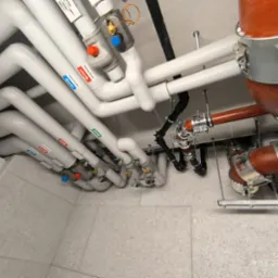 Kompleksowa instalacja grzewcza: rury z oznaczeniami (czerwony/niebieski), zawory, elementy mocujące. Widok z dołu, oświetlenie techniczne.