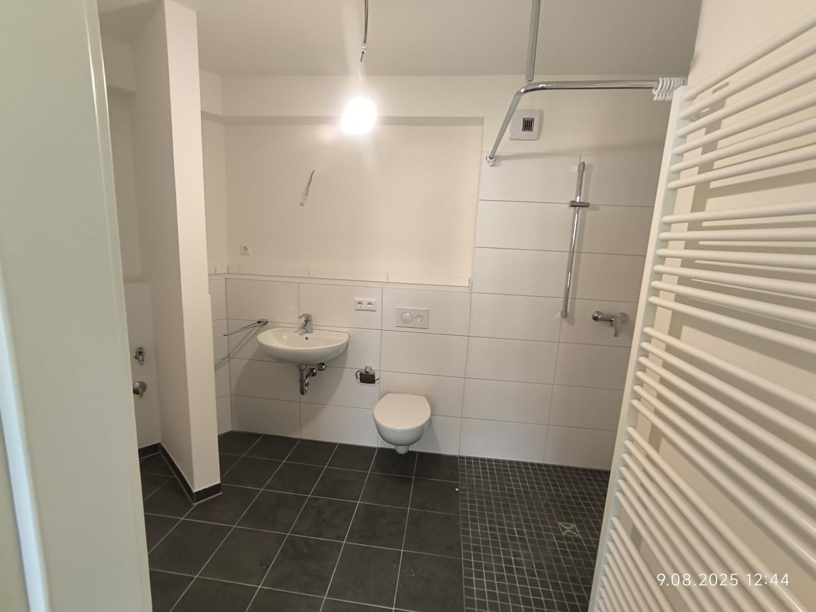 Wykończona łazienka w minimalistycznym stylu: białe płytki, umywalka, toaleta, grzejnik drabinkowy i czarne płytki podłogowe. Widok z otwartych drzwi.