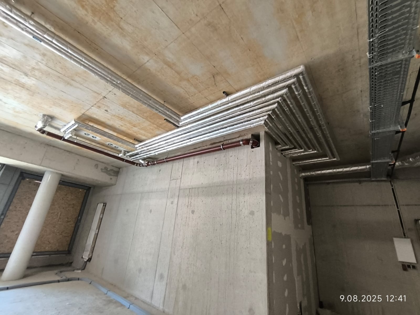 Instalacja rur grzewczych pod sufitem w surowym wnętrzu, zabezpieczone izolacją, widoczne elementy mocujące, surowy beton i okablowanie.