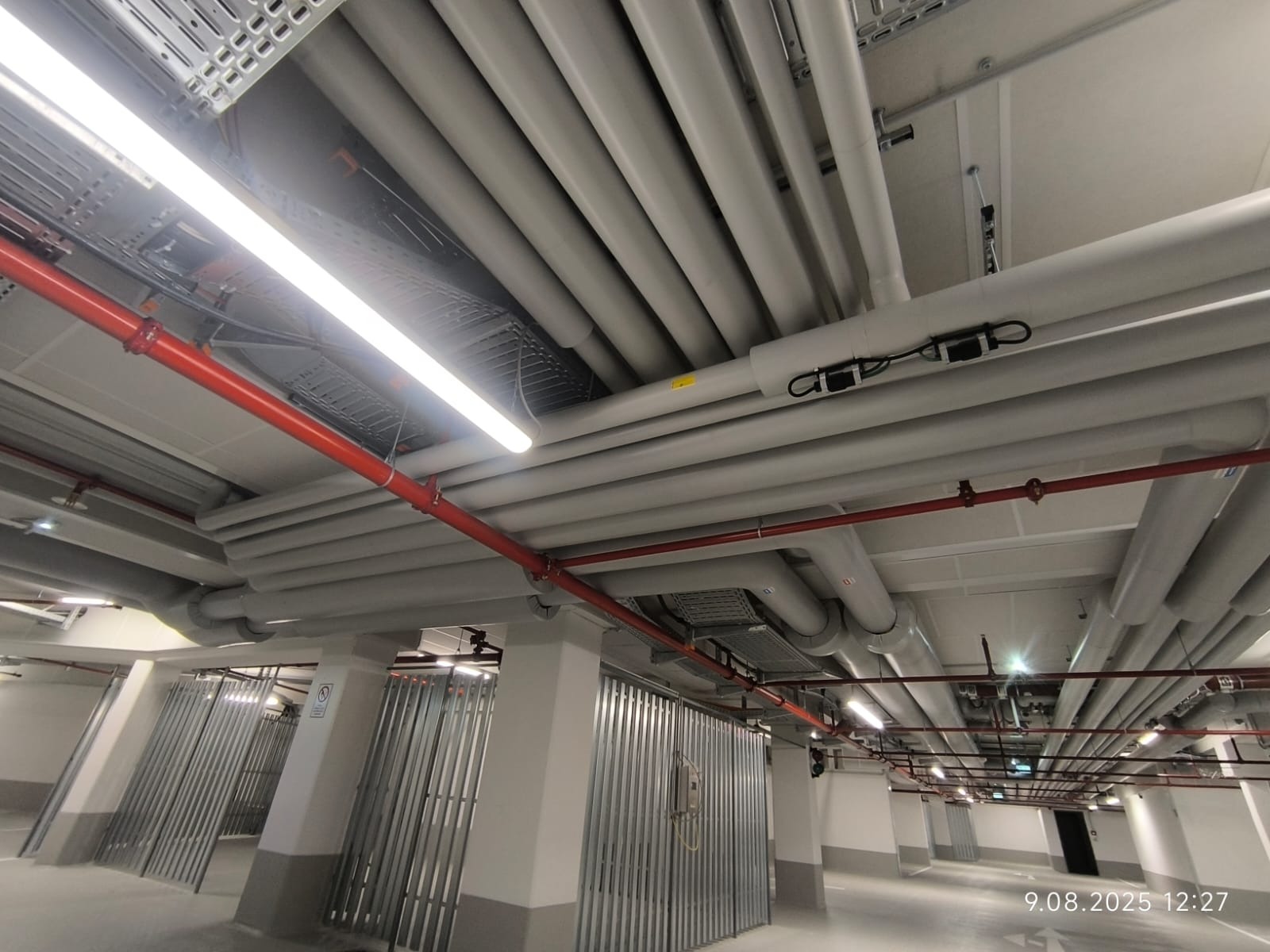 Instalacja rur pod sufitem parkingu podziemnego, widoczne pionowe kolumny, oświetlenie liniowe. Rury w kolorze szarym i czerwonym, estetyczne ułożenie.