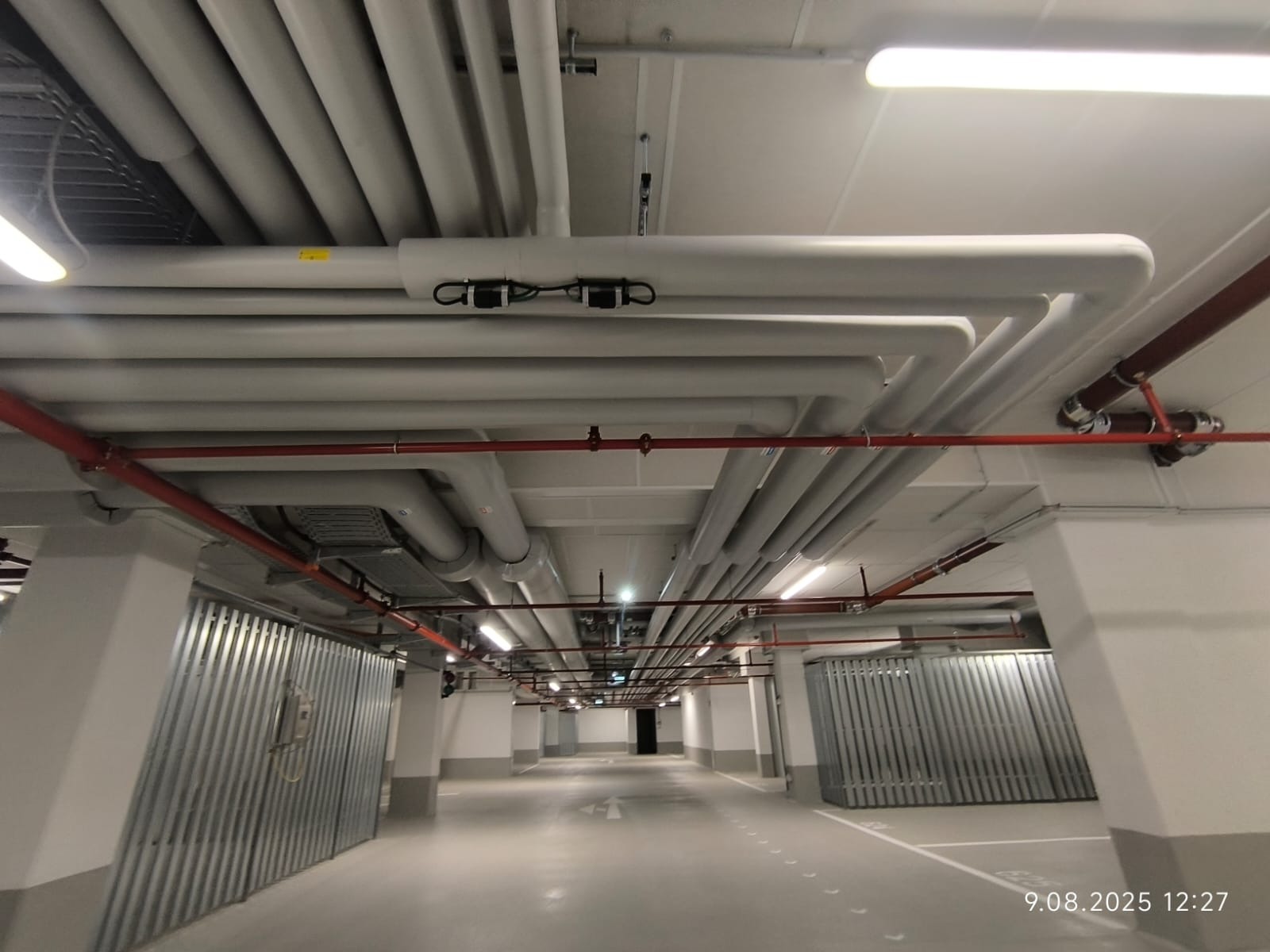 Równoległe rury pod sufitem garażu podziemnego, część rur w kolorze szarym, część czerwonym. Widoczne oświetlenie i elementy instalacji.