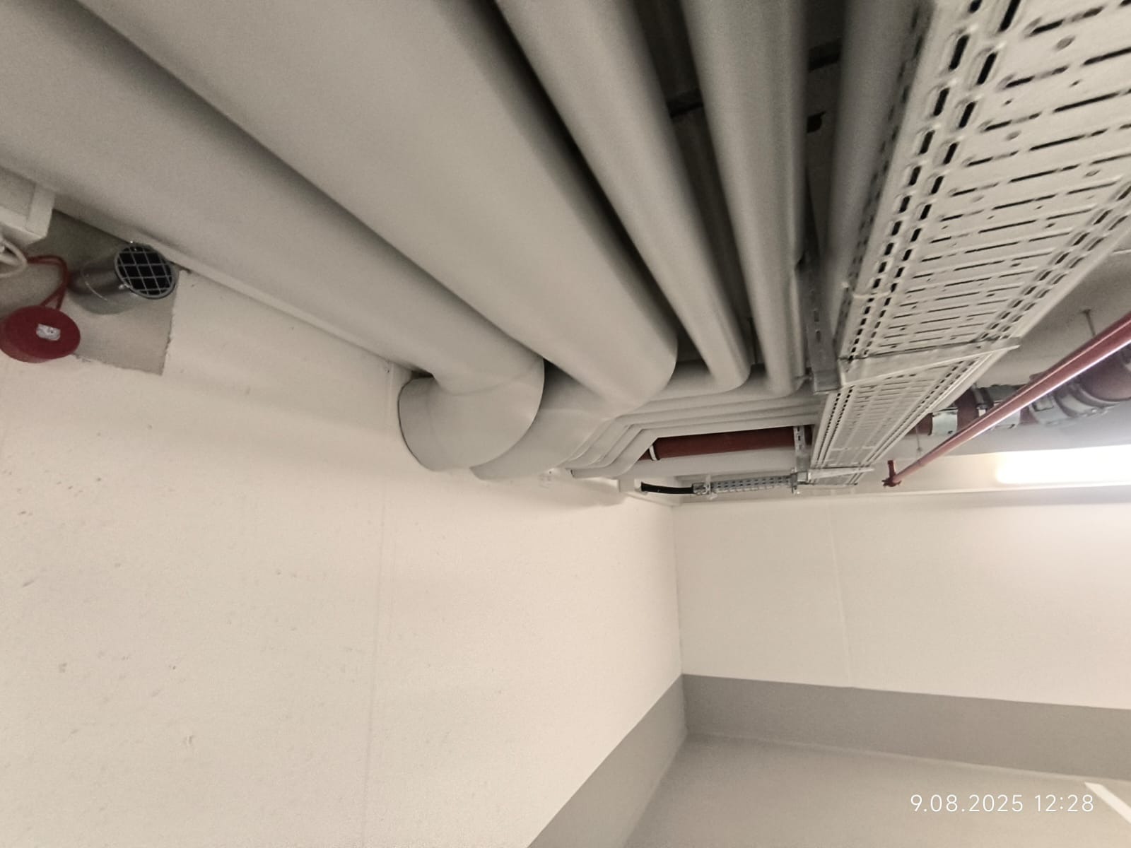 Białe rury instalacyjne pod sufitem w budynku, widoczne także koryta kablowe i element wentylacji. Świeżo pomalowane ściany i cokół.