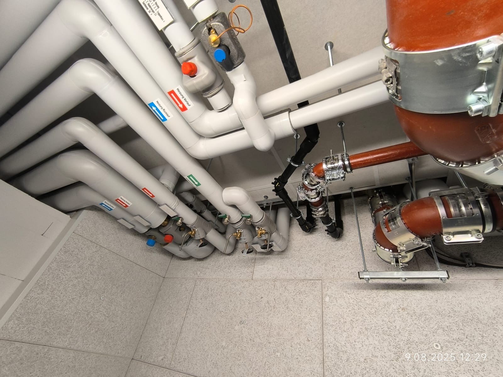 Kompleksowa instalacja grzewcza: rury z oznaczeniami (czerwony/niebieski), zawory, elementy mocujące. Widok z dołu, oświetlenie techniczne.