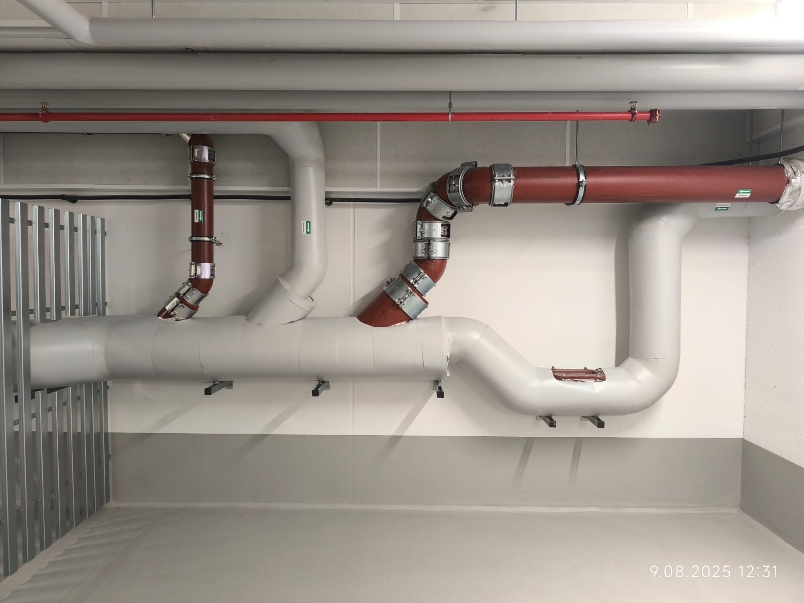 Złożony system rur w piwnicy, z różnymi kolorami i złączkami. Widoczne rury szare, czerwone i brązowe, połączone metalowymi obejmami. Tło stanowi jasna ściana i podłoga.