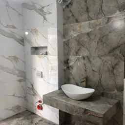 Elegancka łazienka z marmurowymi płytkami w odcieniach bieli i szarości, owalna umywalka na blacie z tego samego materiału, złota bateria, minimalistyczny design.