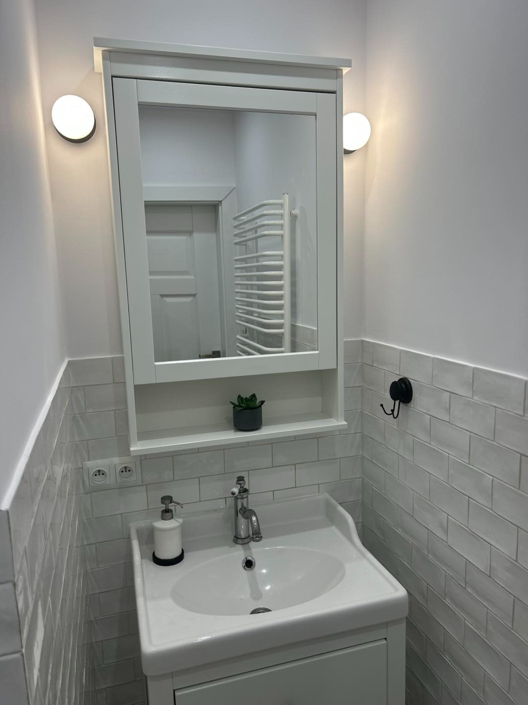 Biała umywalka z szafką i lustrem w ramie, oświetlona kulistymi lampami. Na blacie dozownik mydła, na półce roślina. W tle widoczny grzejnik drabinkowy.