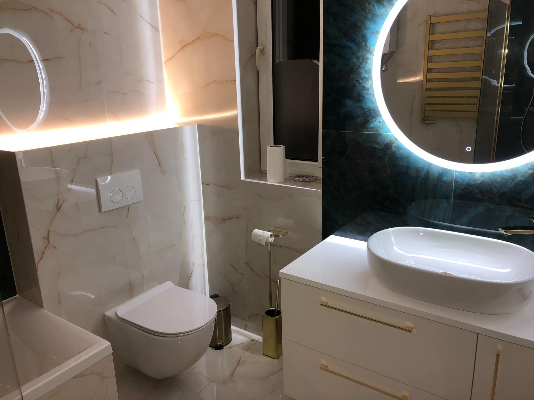 Elegancka łazienka w odcieniach bieli i złota: umywalka nablatowa, toaleta podwieszana i okrągłe lustro z podświetleniem LED. Marmurowe płytki na ścianach.