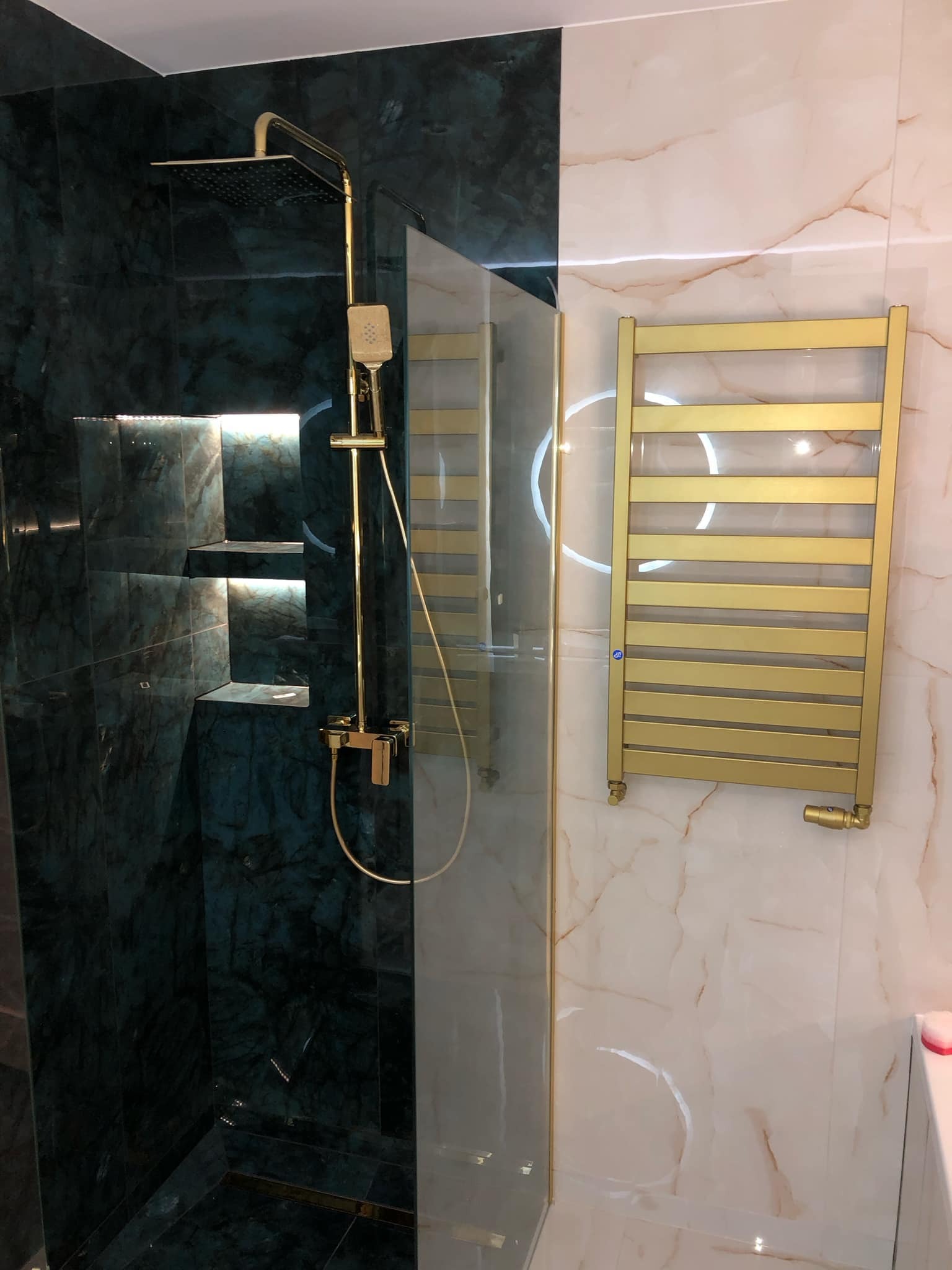 Elegancka łazienka: zielony marmur pod prysznicem z półkami i złotą armaturą, obok złoty grzejnik na ścianie z marmuru w odcieniach beżu.