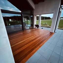 MIĘDZY SĘKAMI JAKUB WIERNICKI-BALCER - Montaż drewnianego tarasu przy nowoczesnym domu z dużymi oknami. Osoba impregnuje deski tarasowe. Widok na ogród i niebo.