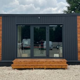 MIĘDZY SĘKAMI JAKUB WIERNICKI-BALCER - Nowoczesna altana z szarymi panelami i drewnianymi akcentami, widoczne drzwi szklane i drewniane schodki wejściowe. Całość na żwirowym podłożu.