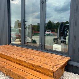 MIĘDZY SĘKAMI JAKUB WIERNICKI-BALCER - Nowe, drewniane schody wejściowe z jasnego drewna prowadzą do szarego budynku z dużymi, przeszklonymi drzwiami. Widoczne odbicia otoczenia w szybach.