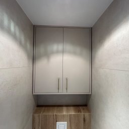 MIĘDZY SĘKAMI JAKUB WIERNICKI-BALCER - Zamknięta szafka wisząca w łazience, z dwoma drzwiczkami i długimi uchwytami, nad zabudową toalety z brązowymi płytkami imitującymi drewno.