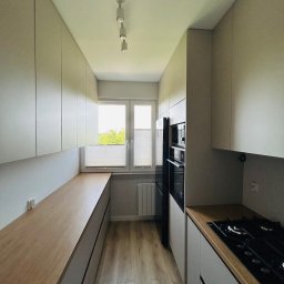 MIĘDZY SĘKAMI JAKUB WIERNICKI-BALCER - Wąska kuchnia z zabudową po obu stronach, jasne fronty szafek i drewniany blat roboczy. Widoczne okno z roletami plisowanymi i sprzęt AGD w zabudowie.