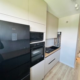 MIĘDZY SĘKAMI JAKUB WIERNICKI-BALCER - Nowoczesna kuchnia w odcieniach beżu i bieli w Wygodzie. Zabudowa kuchenna z czarnym sprzętem AGD i drewnianym blatem. Widoczne piony instalacyjne.