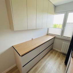 MIĘDZY SĘKAMI JAKUB WIERNICKI-BALCER - Jasna zabudowa meblowa z drewnianym blatem przy oknie, z górnymi szafkami i dolnymi szufladami z czarnymi akcentami, na podłodze panele imitujące drewno.