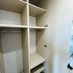 MIĘDZY SĘKAMI JAKUB WIERNICKI-BALCER - Jasna szafa na wymiar z otwartymi drzwiami, widoczne półki i drążki. Dolne półki na buty. Na podłodze leży wiertarka. Minimalistyczny design.