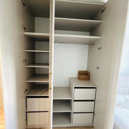 MIĘDZY SĘKAMI JAKUB WIERNICKI-BALCER - Jasna szafa na wymiar z otwartymi drzwiami, ukazująca półki i szuflady. Widoczna kartonowa skrzynka na jednej z półek. Ujęcie z perspektywy wnętrza pomieszczenia.