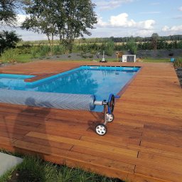 MIĘDZY SĘKAMI JAKUB WIERNICKI-BALCER - Drewniany taras okalający basen z błękitną wodą, częściowo przykryty roletą. W tle trawa, drzewa i krzewy, co tworzy naturalne otoczenie.