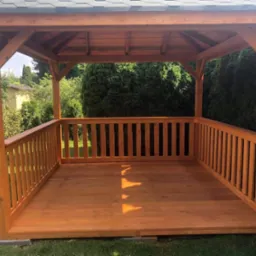 Drewniana altana z balustradą w Wygodzie, zadaszona gontem bitumicznym, otoczona zielenią. Widok z wnętrza, słoneczny dzień.